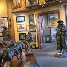 Gallery room with sculptures of wolves, a Native American figure, and a person in western attire, surrounded by paintings of wildlife and western scenes